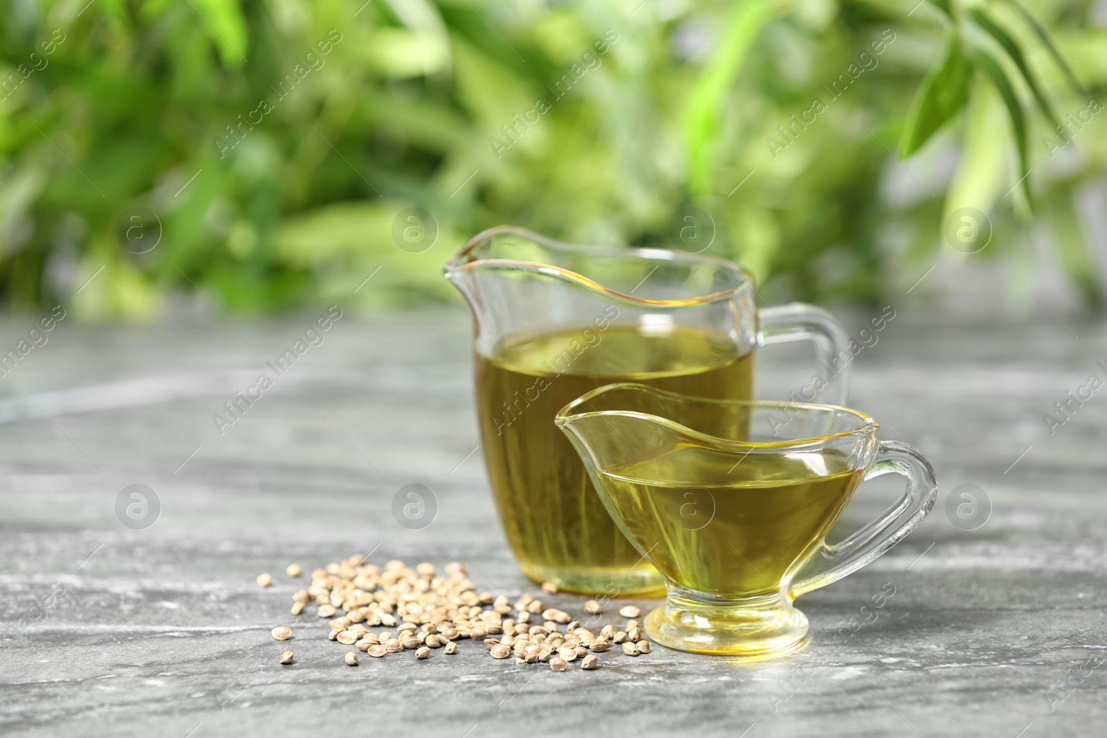 Photo of Glass pitcher and gravy boat with hemp oil on grey table against blurred background