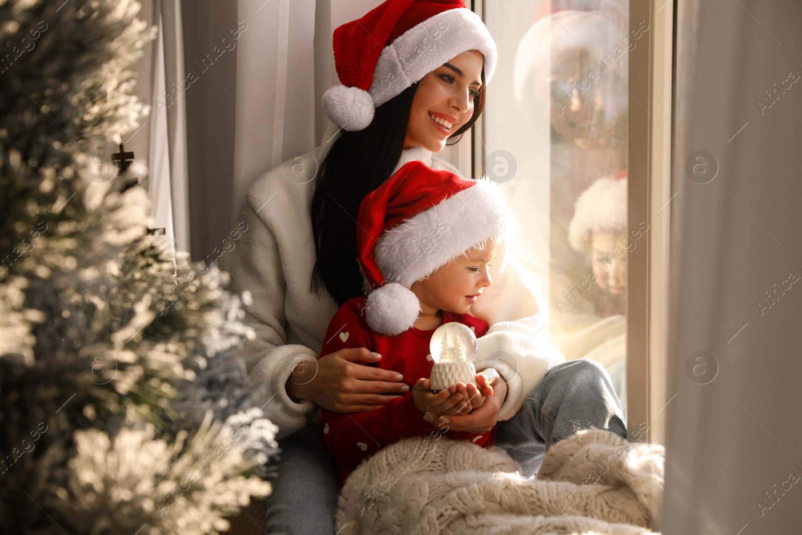 Photo of Mother and daughter in Santa hats playing with snow globe near window