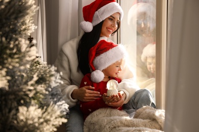 Mother and daughter in Santa hats playing with snow globe near window