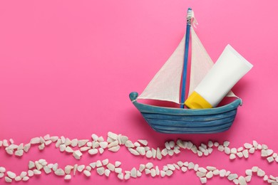 Photo of Suntan cream in toy sailboat and white marble pebbles on pink background, flat lay. Space for text
