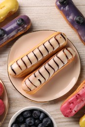 Photo of Different tasty glazed eclairs and blueberries on light wooden table, flat lay