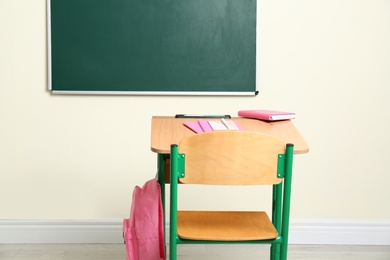 Wooden school desk with stationery and backpack near chalkboard in classroom
