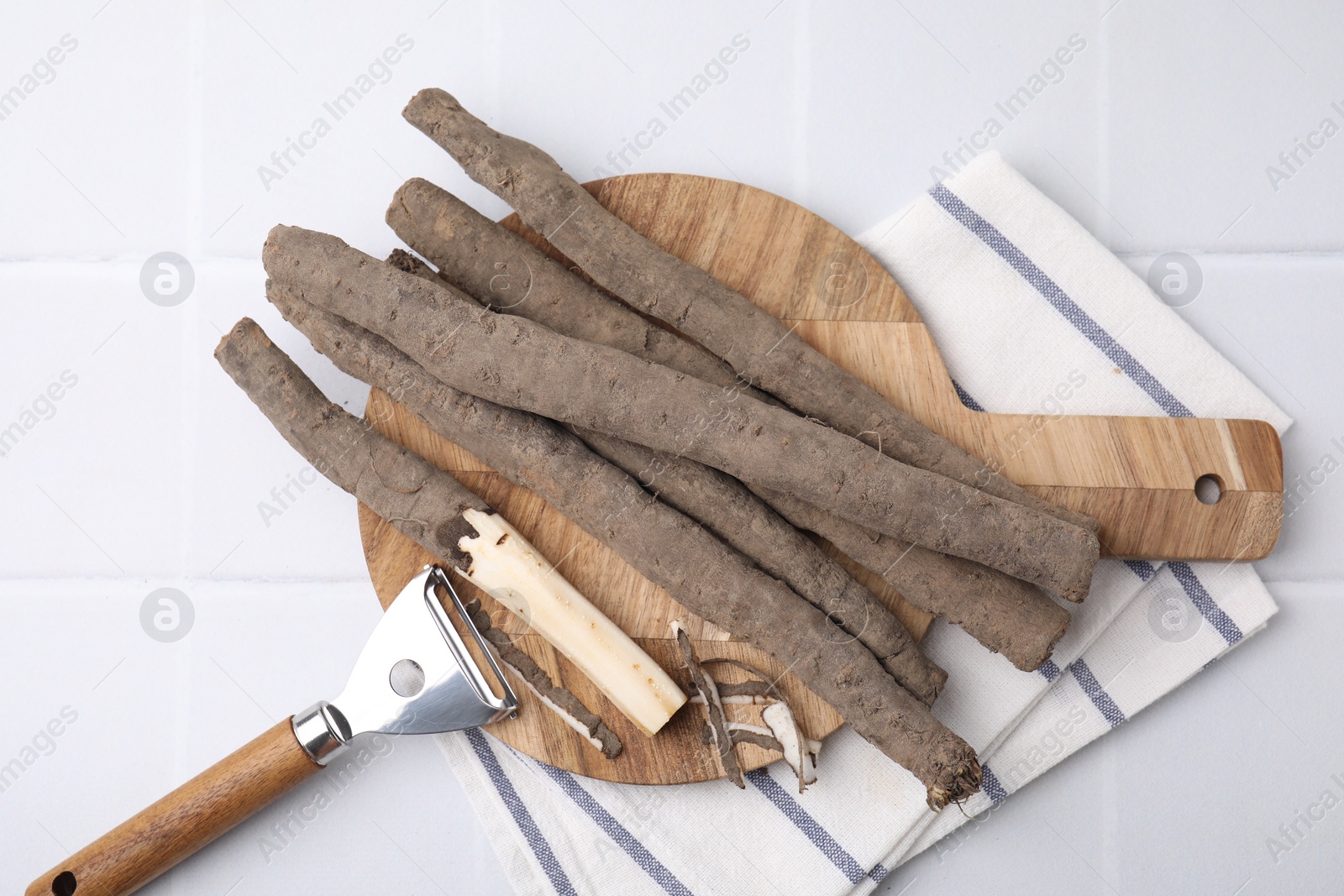 Photo of Raw salsify roots and peeler on white tiled table, flat lay