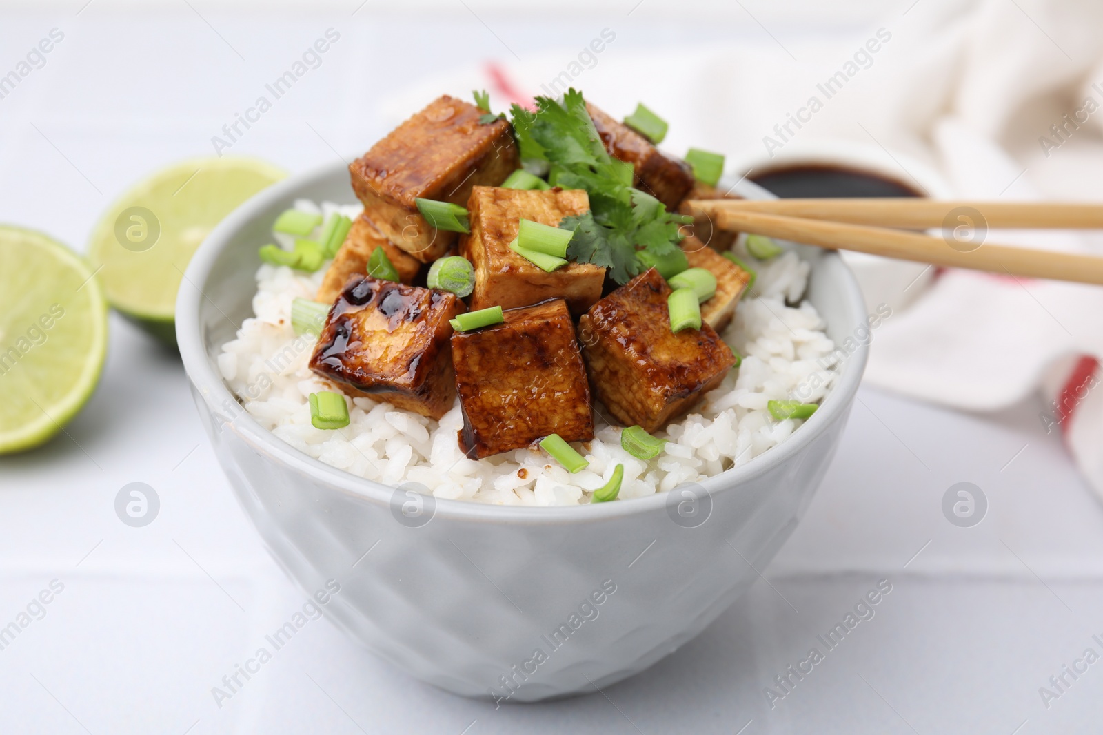 Photo of Bowl of rice with fried tofu and greens on white tiled table, closeup