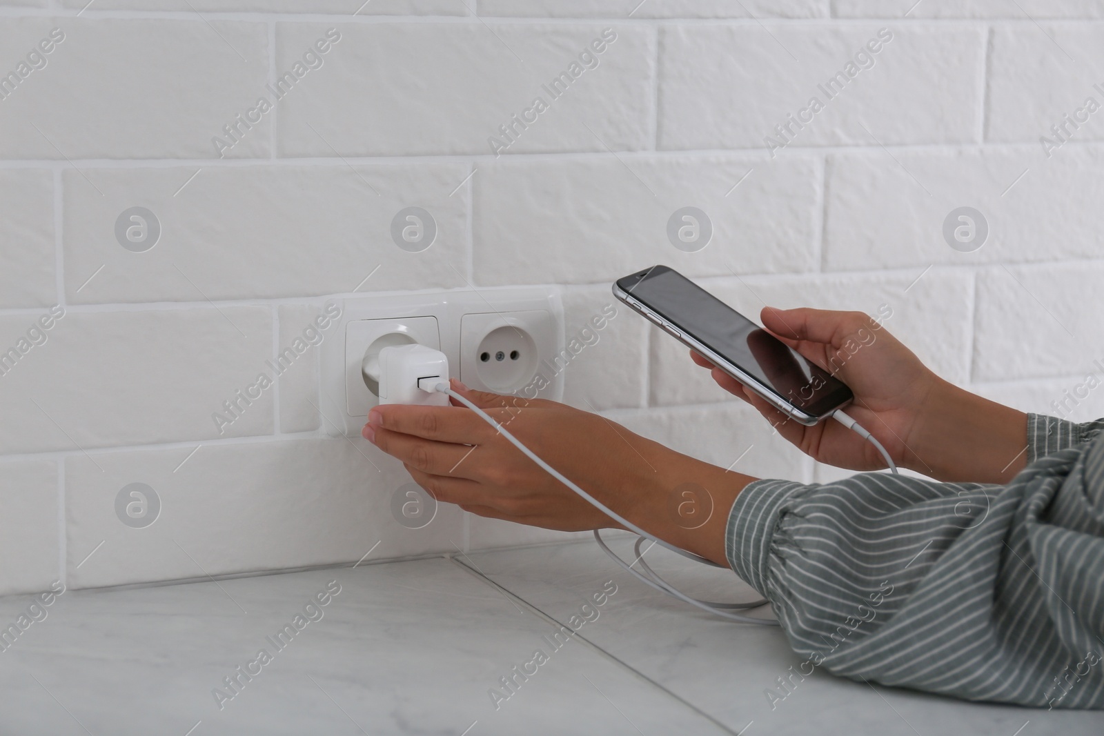 Photo of Woman with mobile phone plugging charger in socket on white brick wall, closeup
