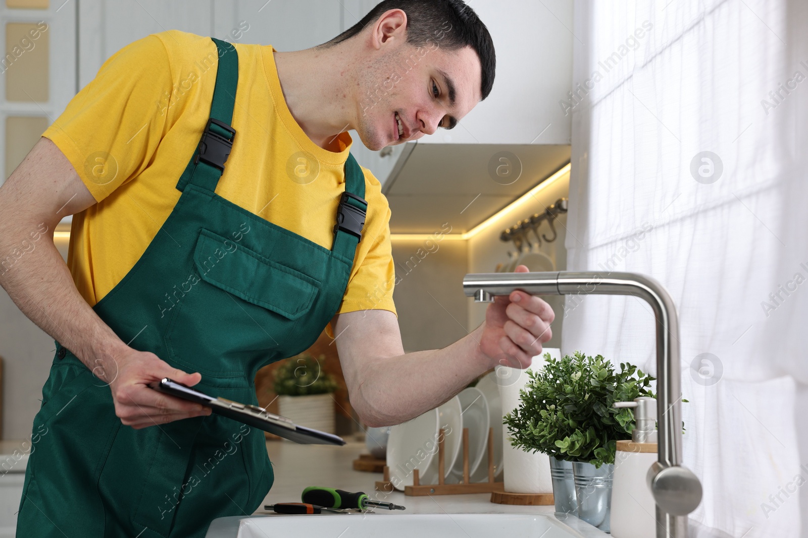 Photo of Smiling plumber with clipboard examining faucet in kitchen