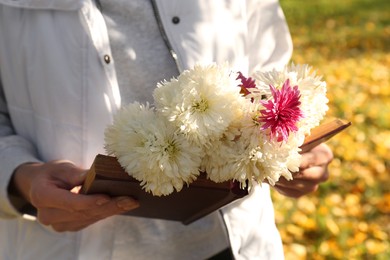 Photo of Woman reading book and holding beautiful flowers outdoors on autumn day, closeup