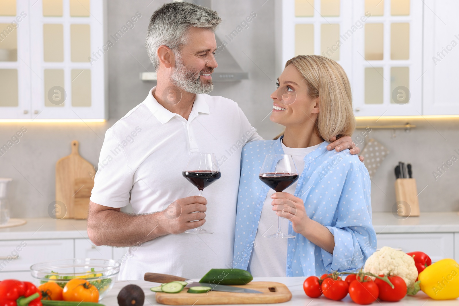 Photo of Happy affectionate couple with glasses of wine cooking together at white table in kitchen