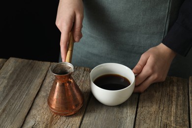 Photo of Turkish coffee. Woman with cezve and cup of freshly brewed beverage at wooden table against black background, closeup