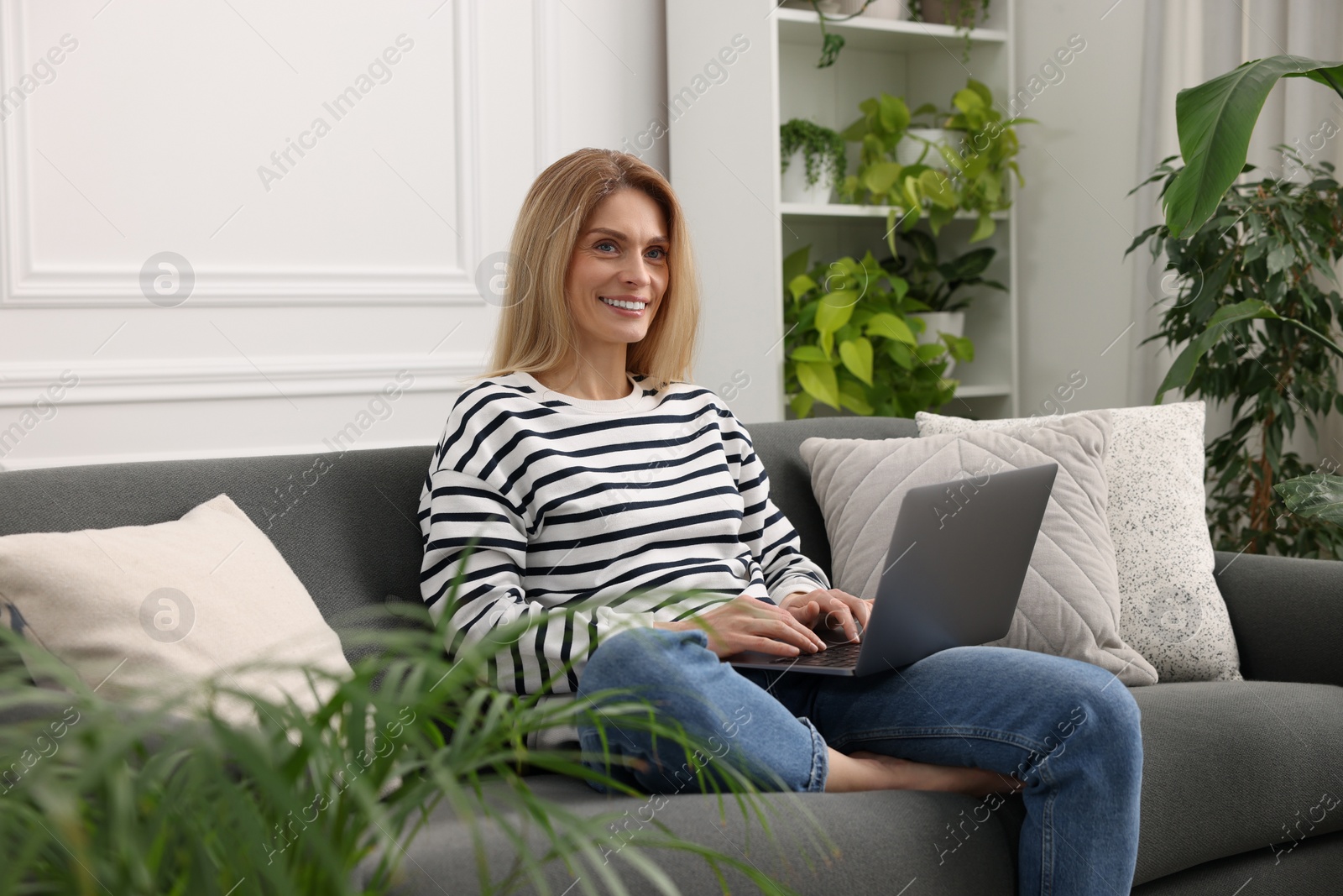 Photo of Woman working with laptop on sofa surrounded by beautiful potted houseplants at home