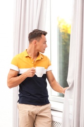 Young man opening window curtains at home