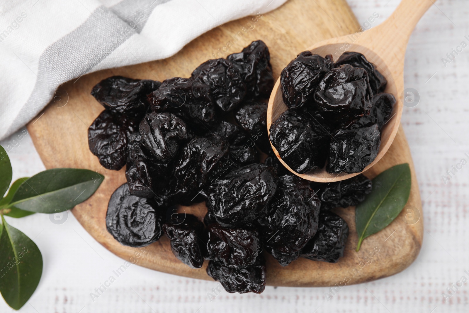 Photo of Spoon with tasty dried prunes and green leaves on light wooden table, top view