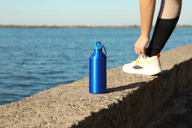 Young woman tying shoelaces near bottle of water at riverside on sunny day