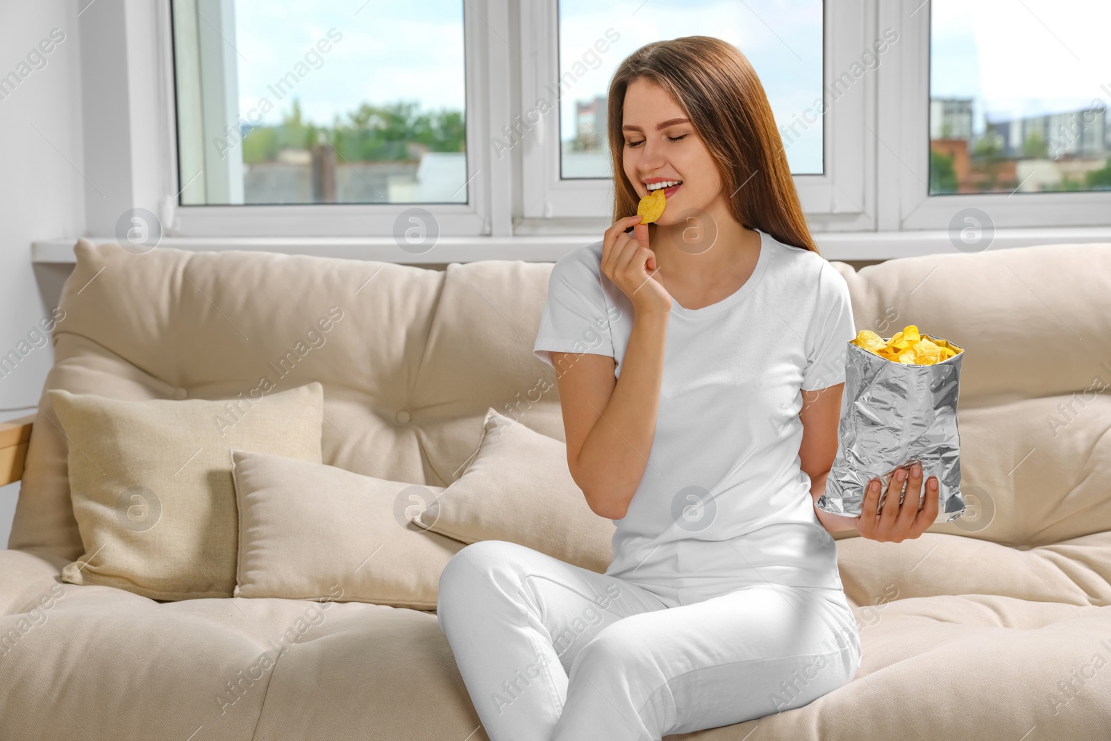 Photo of Pretty young woman eating tasty potato chips on sofa at home