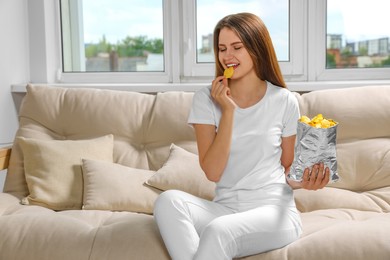 Pretty young woman eating tasty potato chips on sofa at home