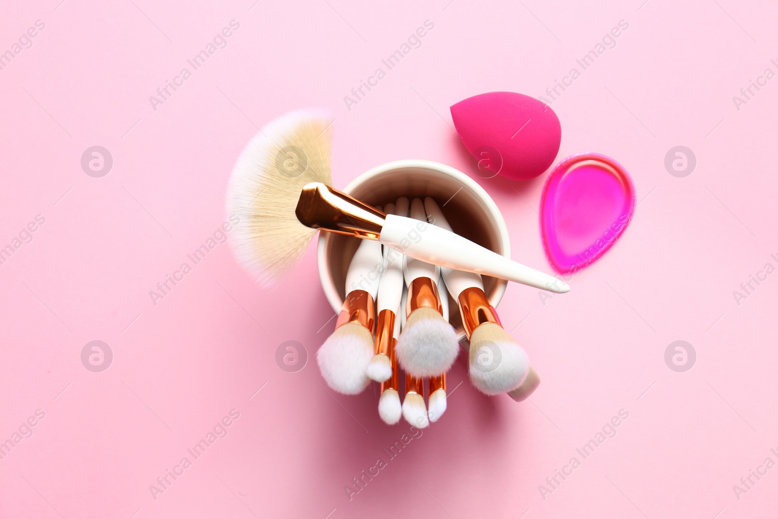 Photo of Holder with professional makeup brushes and sponges on pink background, top view