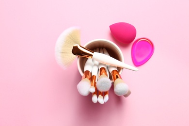 Holder with professional makeup brushes and sponges on pink background, top view