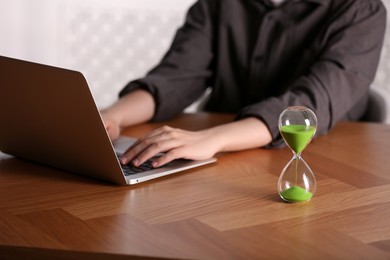 Hourglass with flowing sand on wooden table, selective focus. Man using laptop indoors
