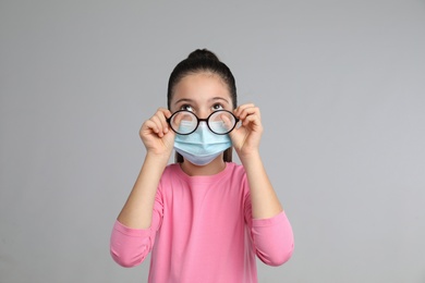 Photo of Little girl wiping foggy glasses caused by wearing medical face mask on grey background. Protective measure during coronavirus pandemic