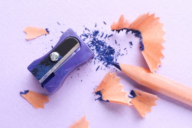 Photo of Blue pencil, sharpener and shavings on pale violet background, flat lay
