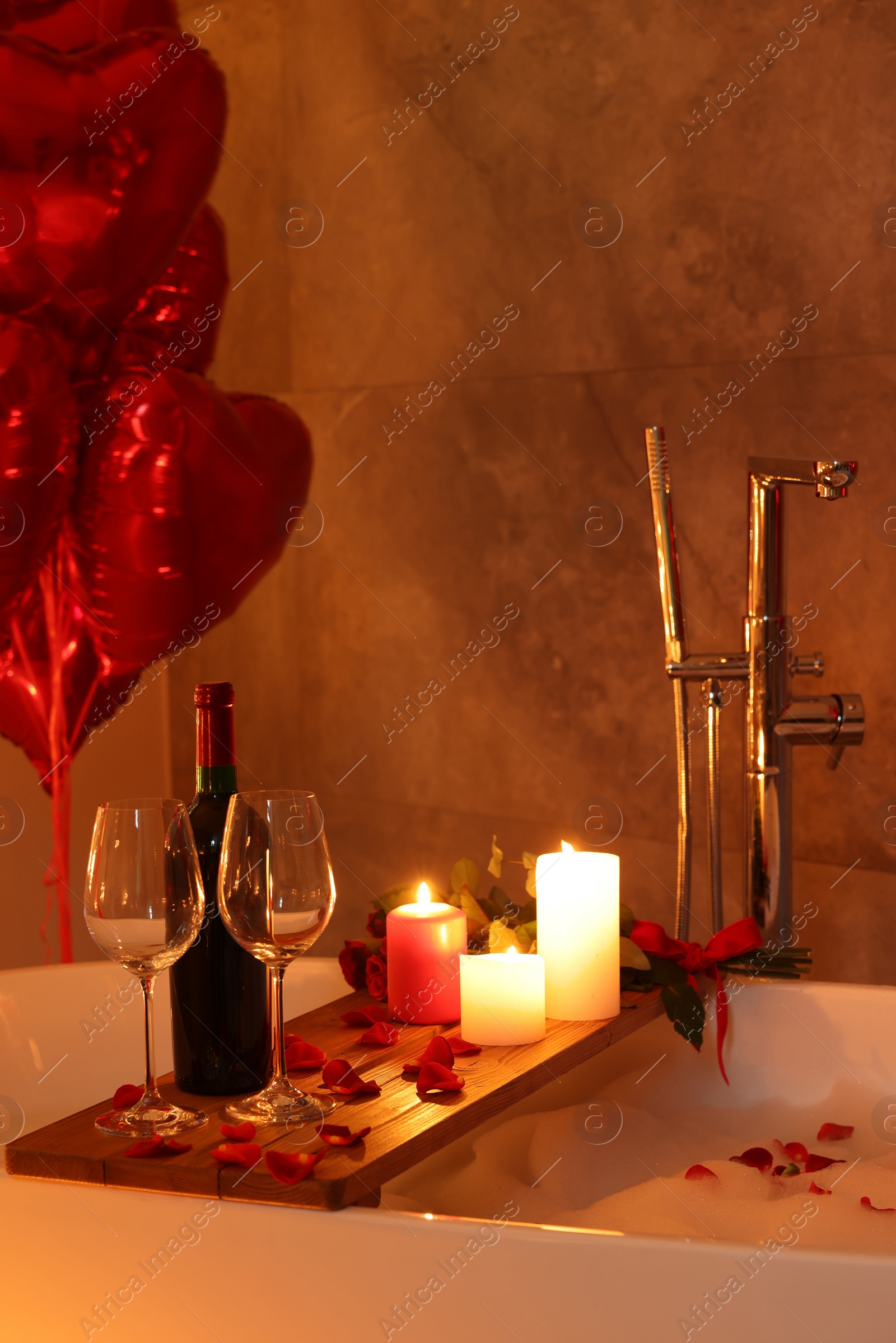 Photo of Wooden tray with wine, burning candles and rose petals on tub in bathroom. Valentine's day celebration