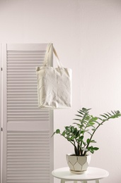 Photo of Eco bag on folding screen near table with houseplant against white wall. Space for design