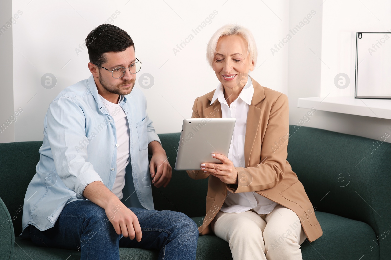 Photo of Happy boss with tablet and employee discussing work issues on sofa in office