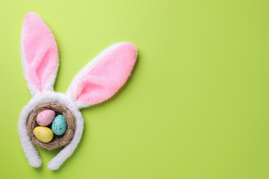 Photo of Headband with Easter bunny ears and dyed eggs in nest on green background, flat lay. Space for text