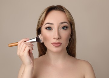 Beautiful young woman applying face powder with brush on grey background