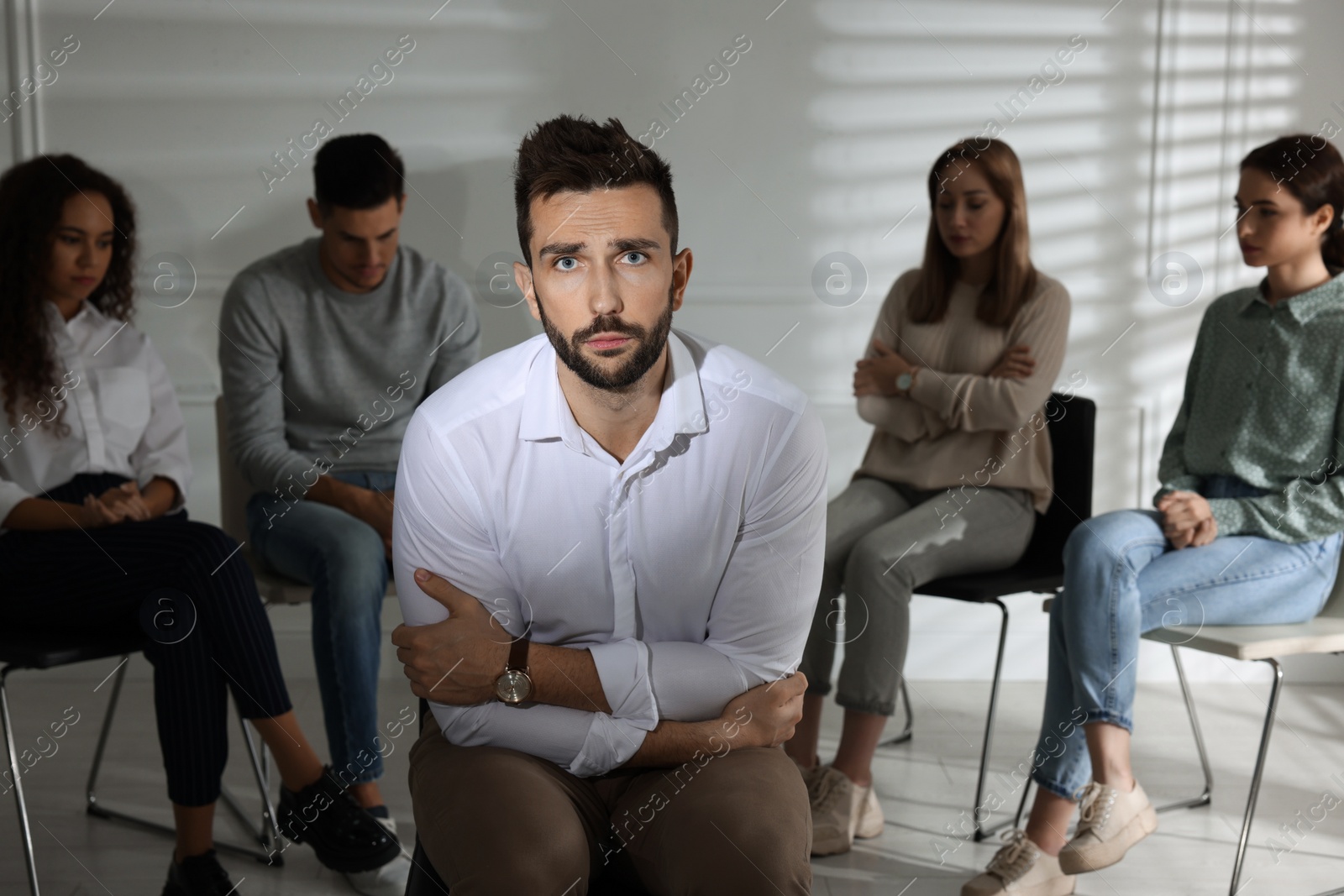 Photo of Unhappy man and group of people behind his back indoors. Therapy session