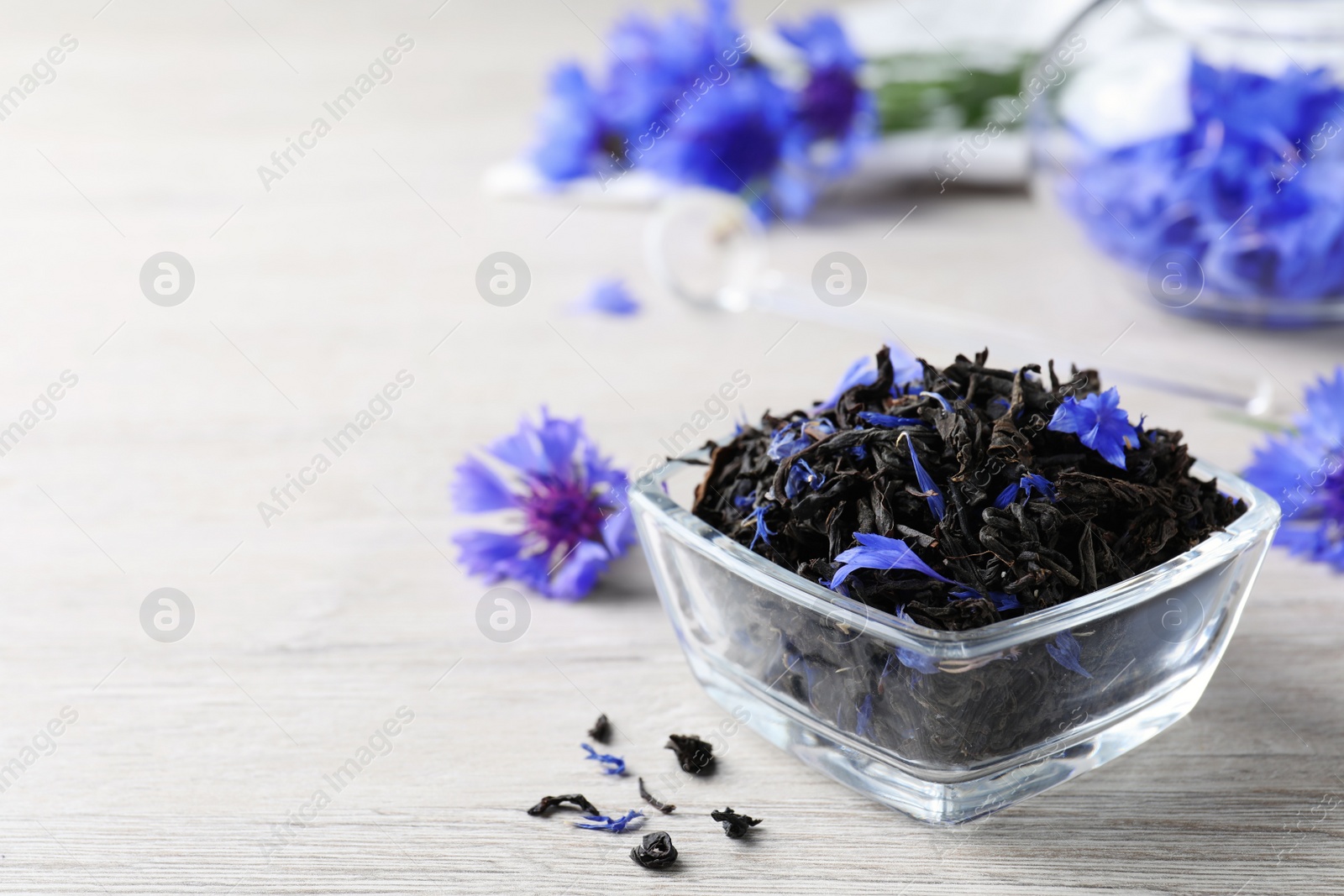 Photo of Glass bowl with dry tea leaves and cornflowers on white wooden table, closeup. Space for text