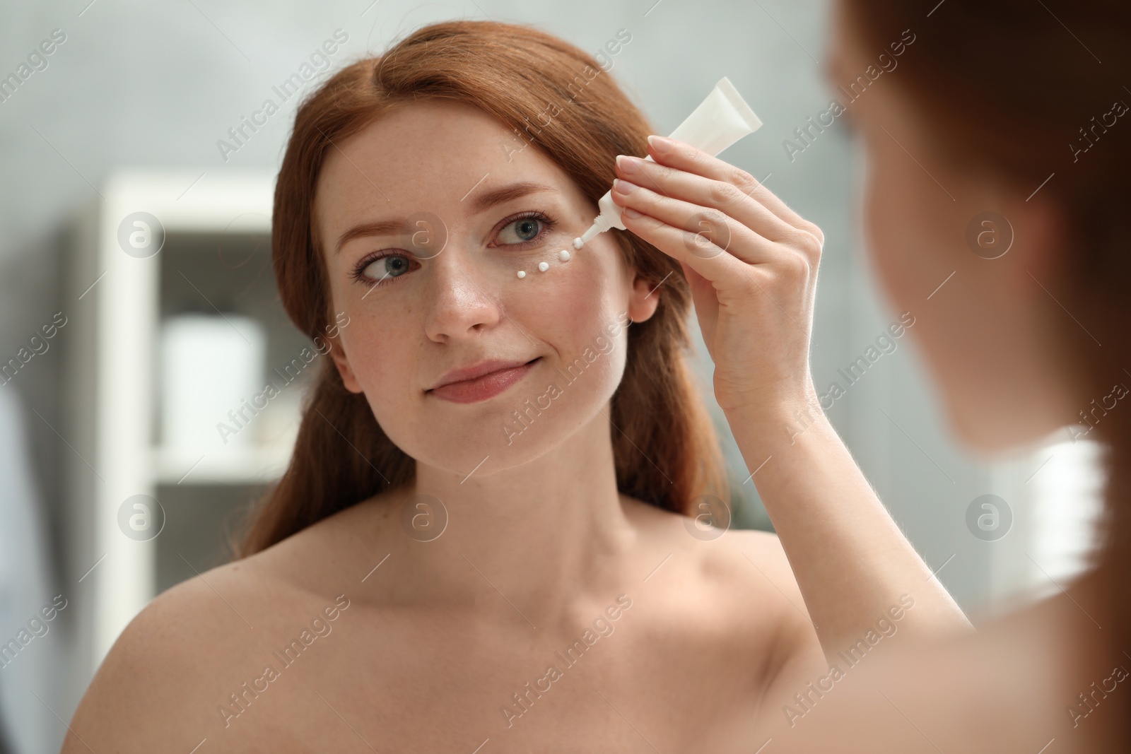 Photo of Beautiful woman with freckles applying cream onto her face near mirror in bathroom