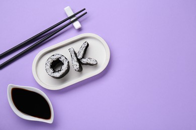 Photo of Word Ok made of sushi rolls, chopsticks and soy sauce on violet background, flat lay. Space for text