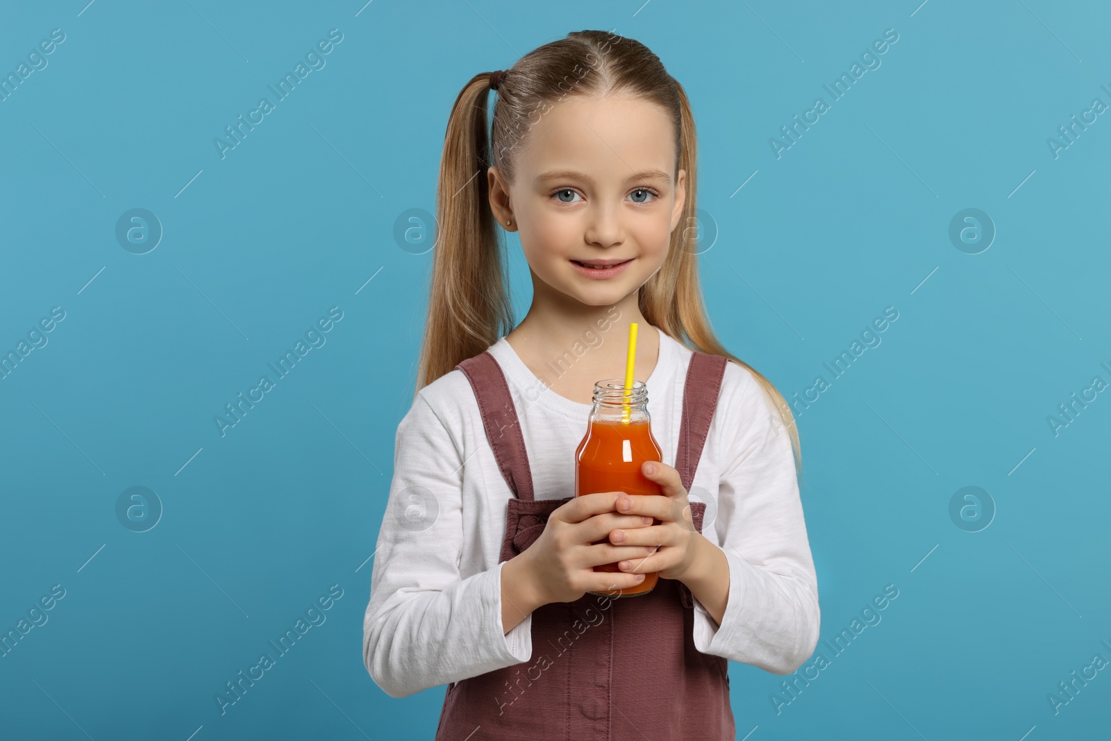 Photo of Cute little girl holding glass bottle of fresh juice with straw on light blue background