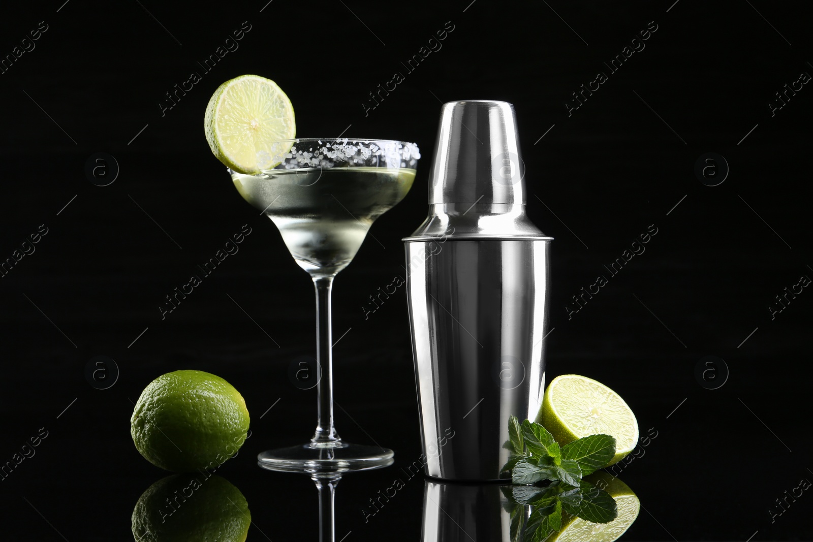 Photo of Metal shaker, delicious cocktail, limes and mint on black mirror surface