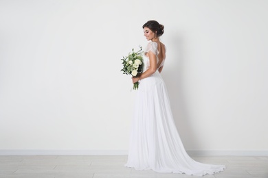 Young bride wearing wedding dress with beautiful bouquet near light wall. Space for text