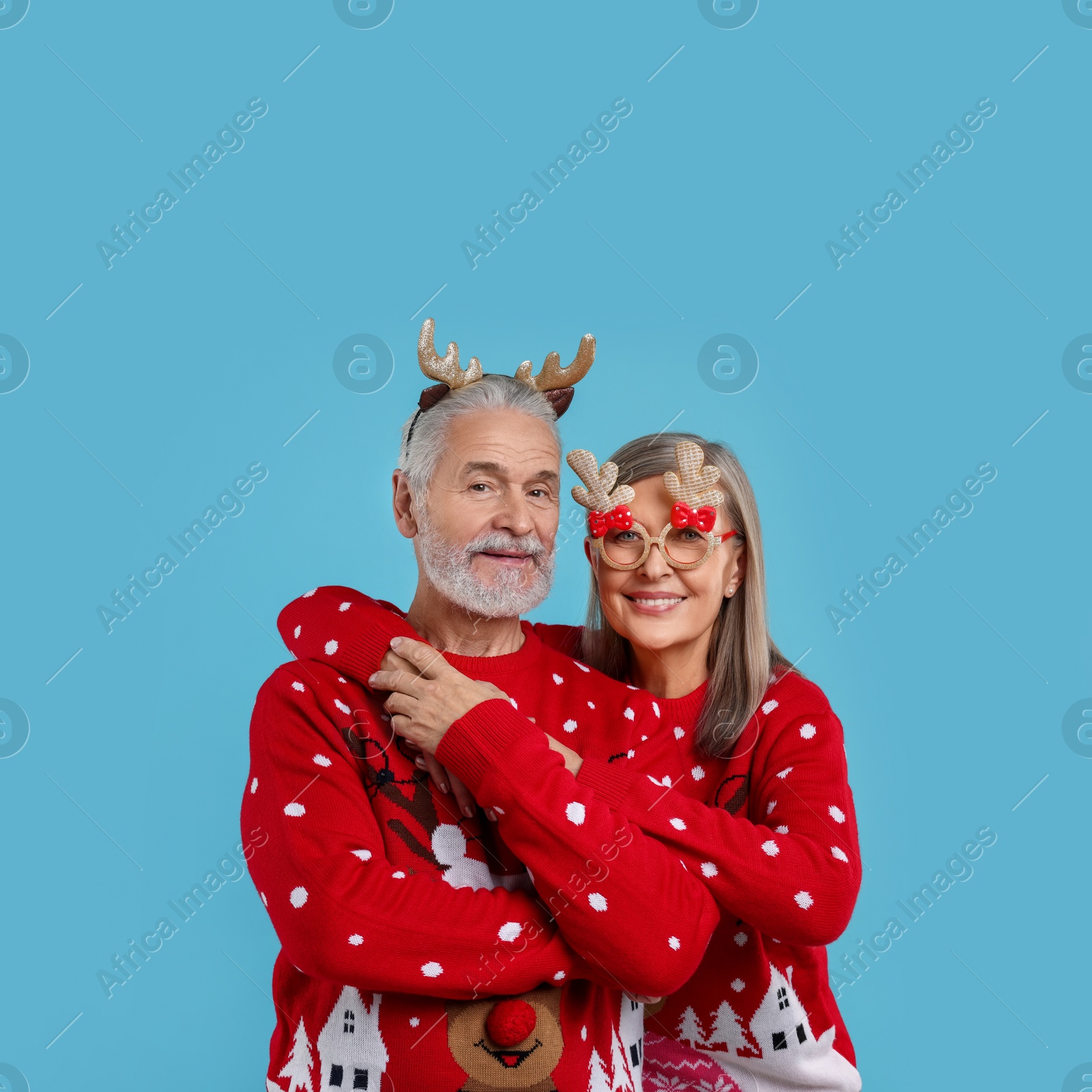 Photo of Senior couple in Christmas sweaters, reindeer headband and funny glasses on light blue background. Space for text