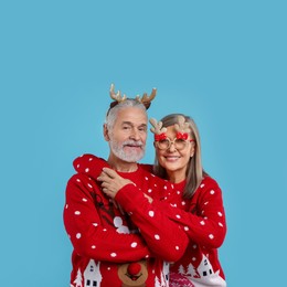 Photo of Senior couple in Christmas sweaters, reindeer headband and funny glasses on light blue background. Space for text