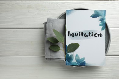 Photo of Card with word Invitation, towel and green leaves on white wooden table, top view. Space for text