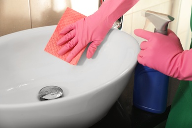 Photo of Woman in protective gloves cleaning bathroom sink with rag, closeup