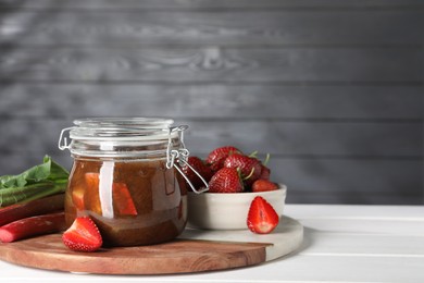 Jar of tasty rhubarb jam, fresh stems and strawberries on white wooden table. Space for text