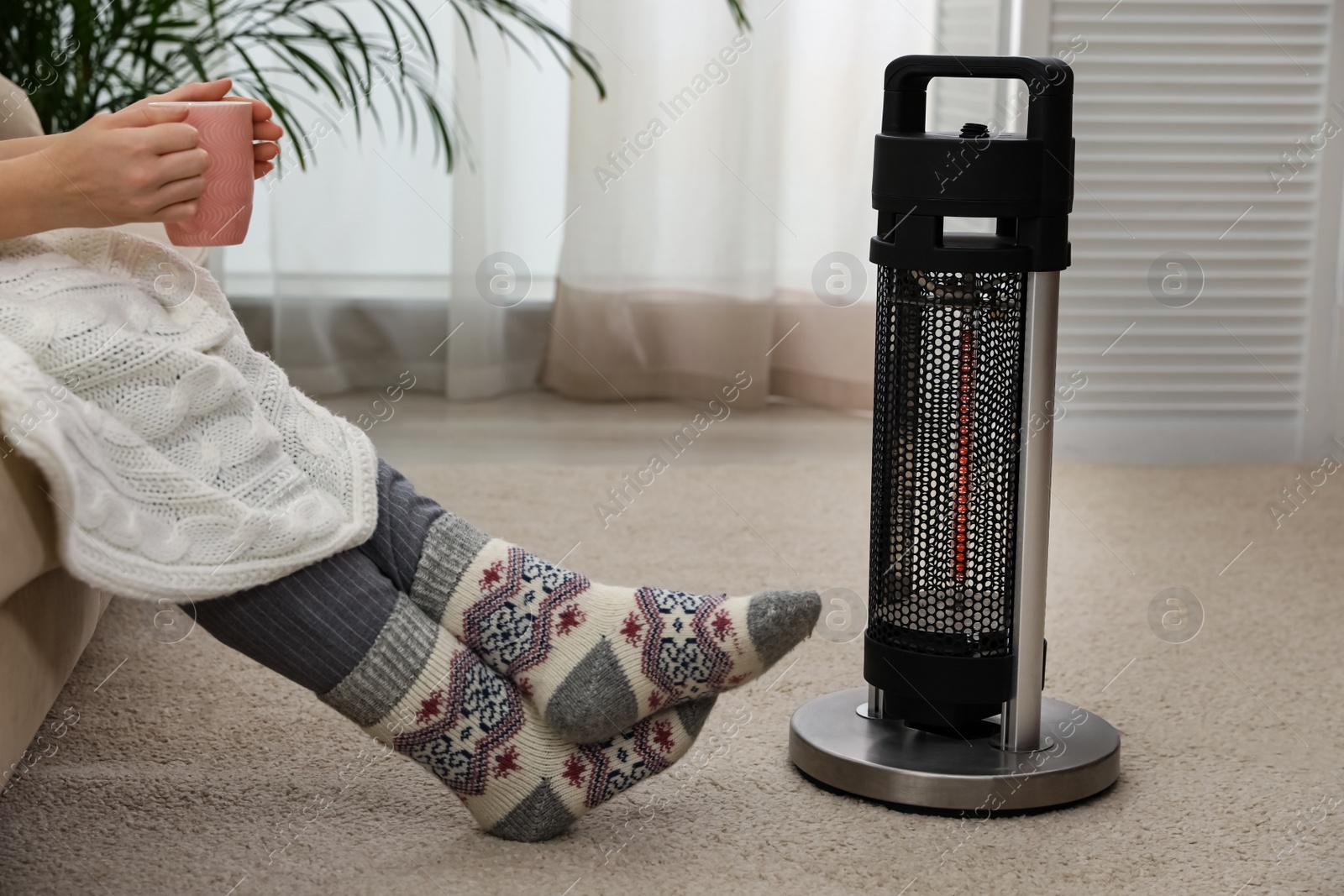 Photo of Woman with tea warming legs near halogen heater at home, closeup