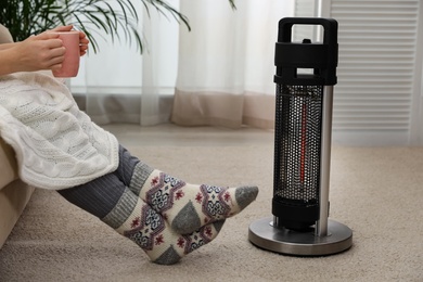 Photo of Woman with tea warming legs near halogen heater at home, closeup