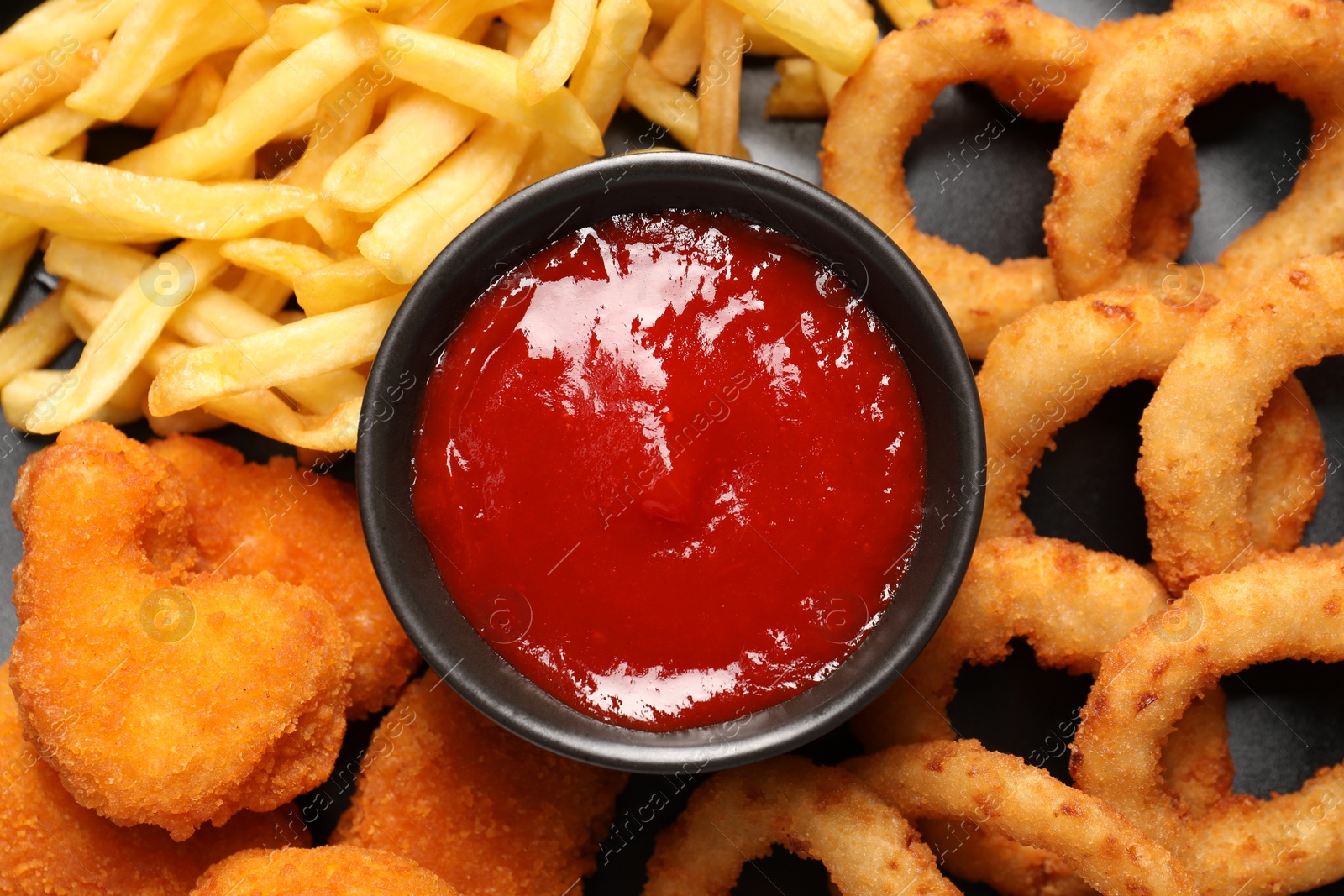 Photo of Bowl with tomato ketchup and different snacks on dark table, closeup