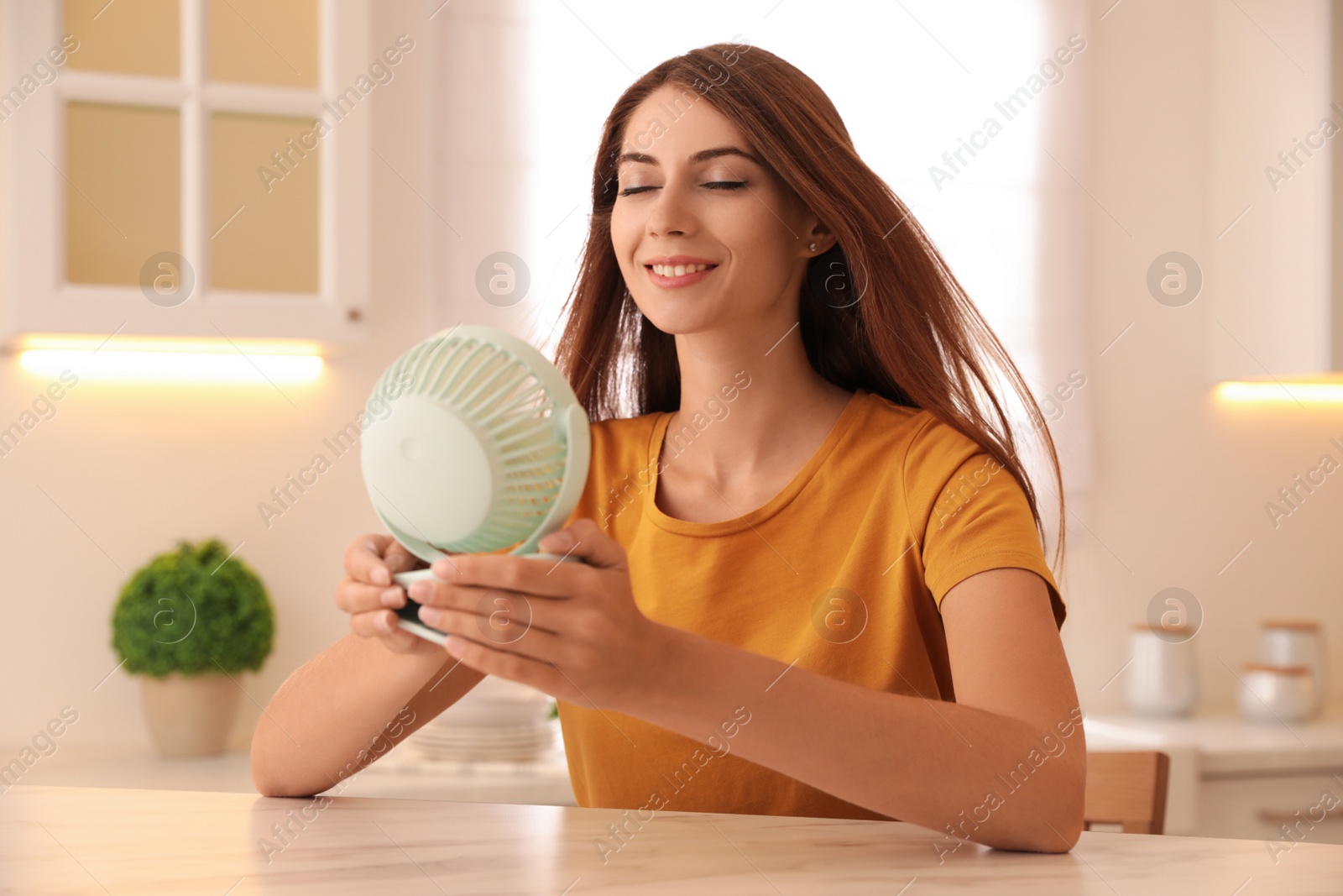 Photo of Woman enjoying air flow from portable fan at table in kitchen. Summer heat