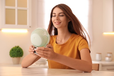 Photo of Woman enjoying air flow from portable fan at table in kitchen. Summer heat