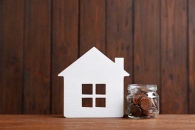 Photo of House model and jar with coins on table against wooden background. Space for text