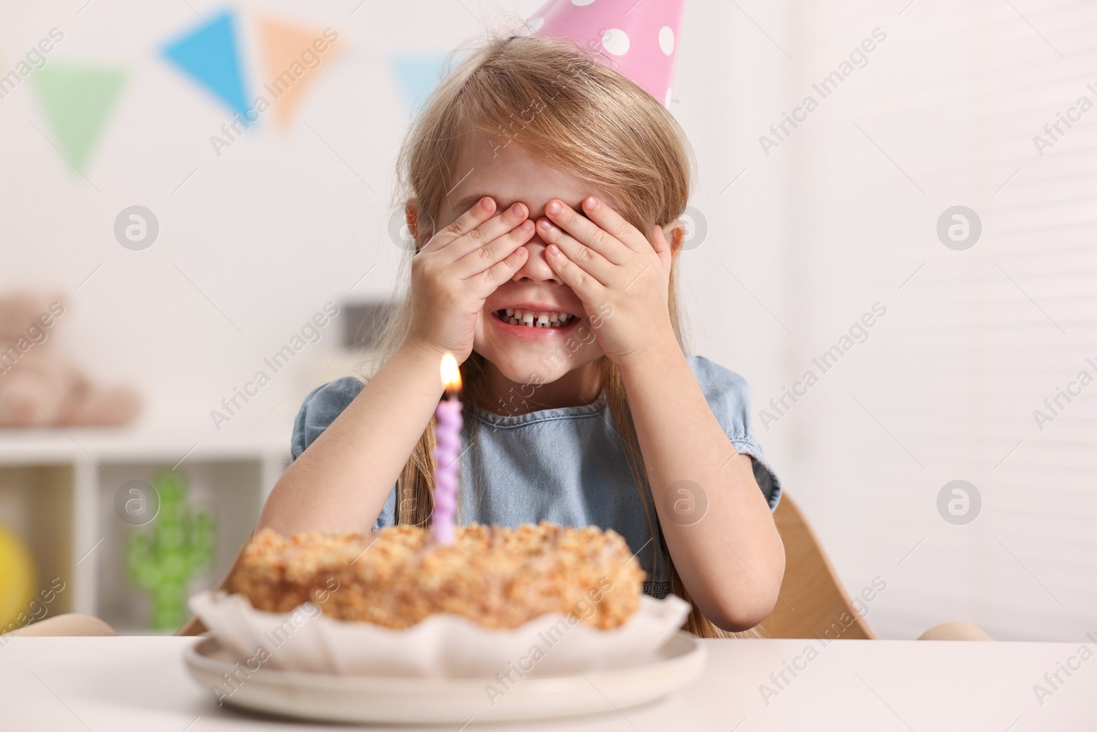 Photo of Cute girl in party hat with birthday cake at table indoors