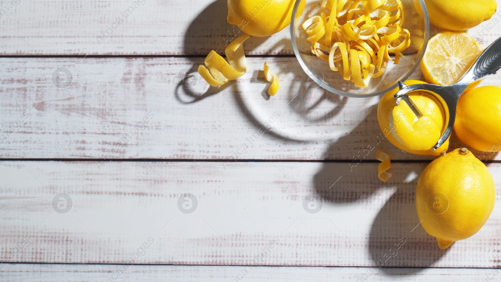 Photo of Bowl with peel pieces, fresh lemons and zester on white wooden table, flat lay. Space for text