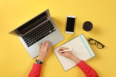 Photo of Woman working with modern laptop and writing in notebook at color table, top view